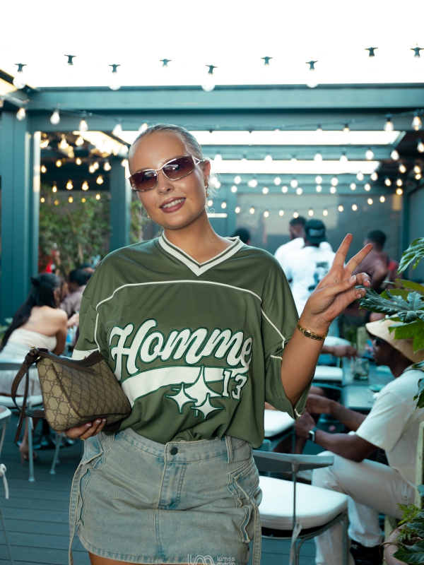 A Woman In A Green Homme Jersey And Denim Skirt Posing At An Outdoor Evening Venue With String Lights.